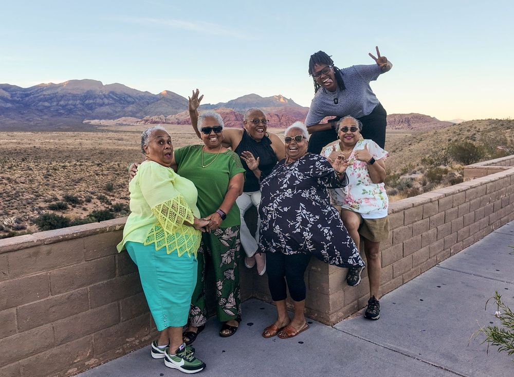 Group of six friends posing and smiling outdoors near Red Rock Canyon desert landscape at sunset.