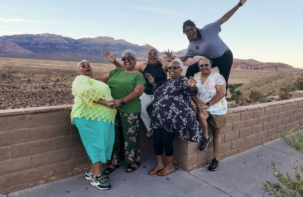 Group of six women posing and smiling in front of a desert mountain landscape at dusk.