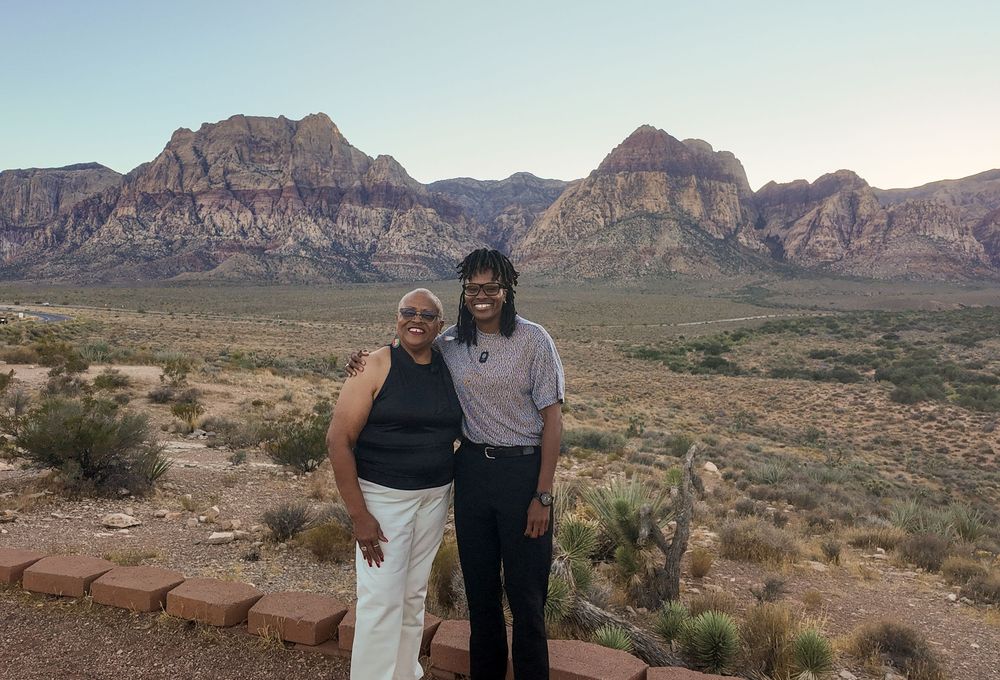 Two women smiling at Red Rock Canyon overlook with red sandstone mountains and desert landscape in background.