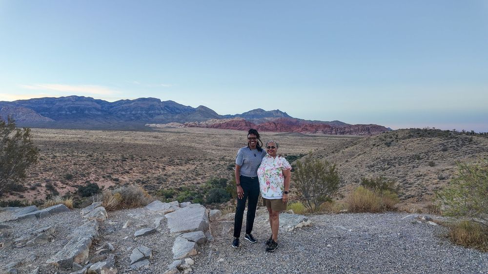 Two people posing at a scenic Red Rock Canyon overlook with desert landscape and mountains at dusk.