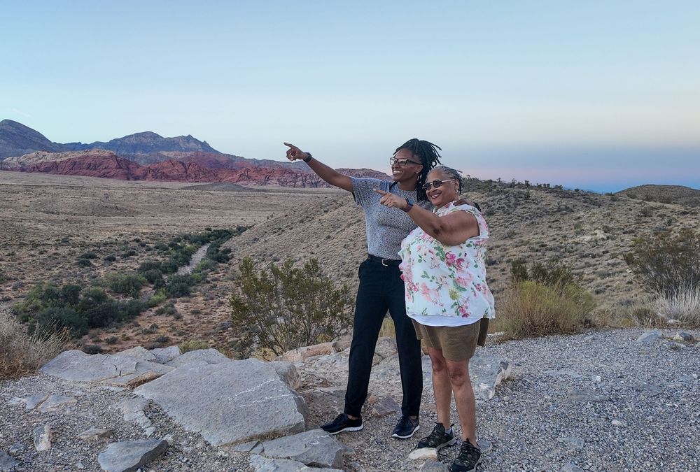 Two women pointing at Red Rock Canyon landscape at dusk, with colorful desert mountains in background.