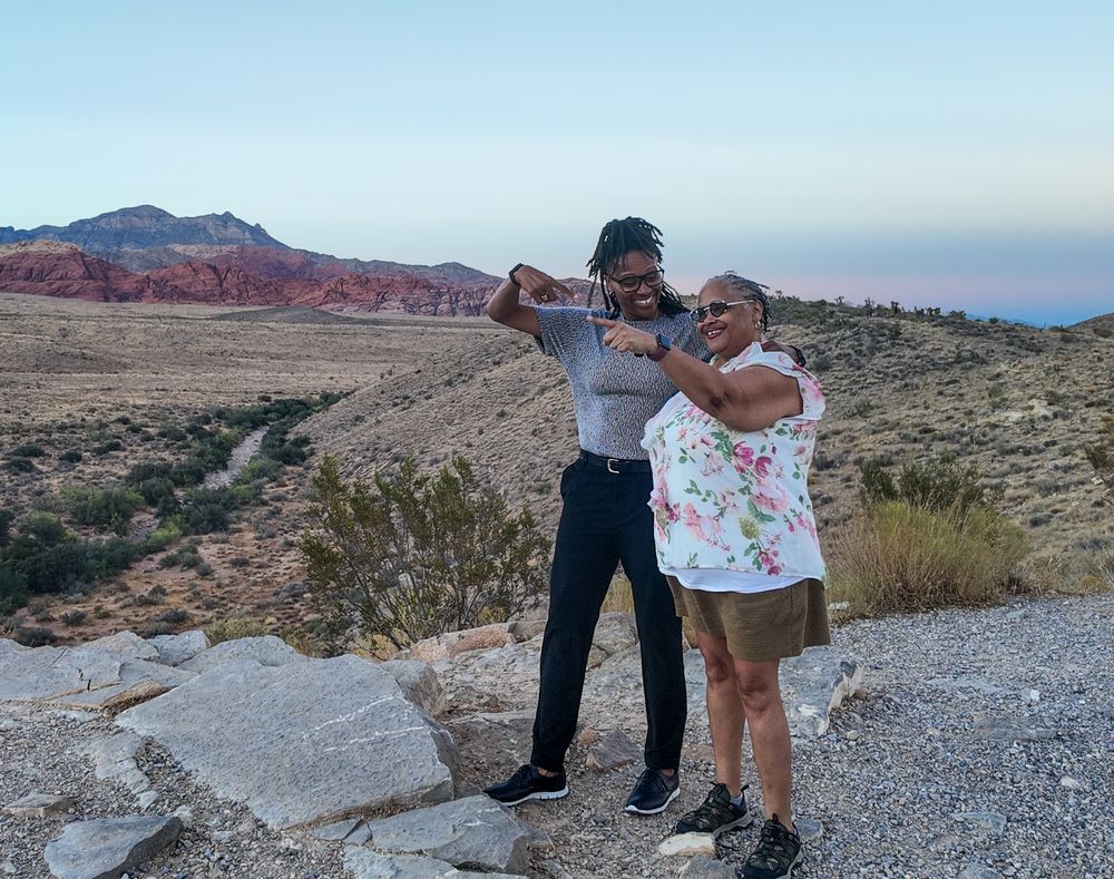 Two people posing and flexing at Red Rock Canyon, Nevada, with scenic red rock formations at sunset.
