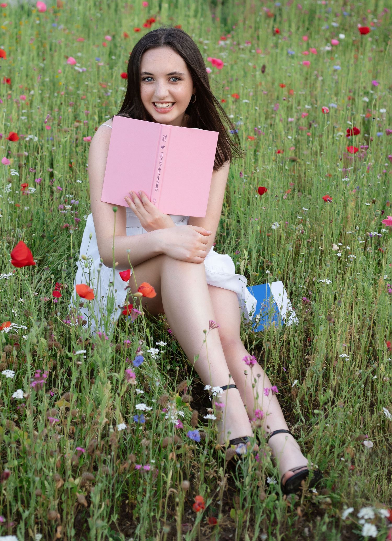 Young woman sitting in a wildflower meadow holding a pink book, wearing a white dress and heeled sandals.