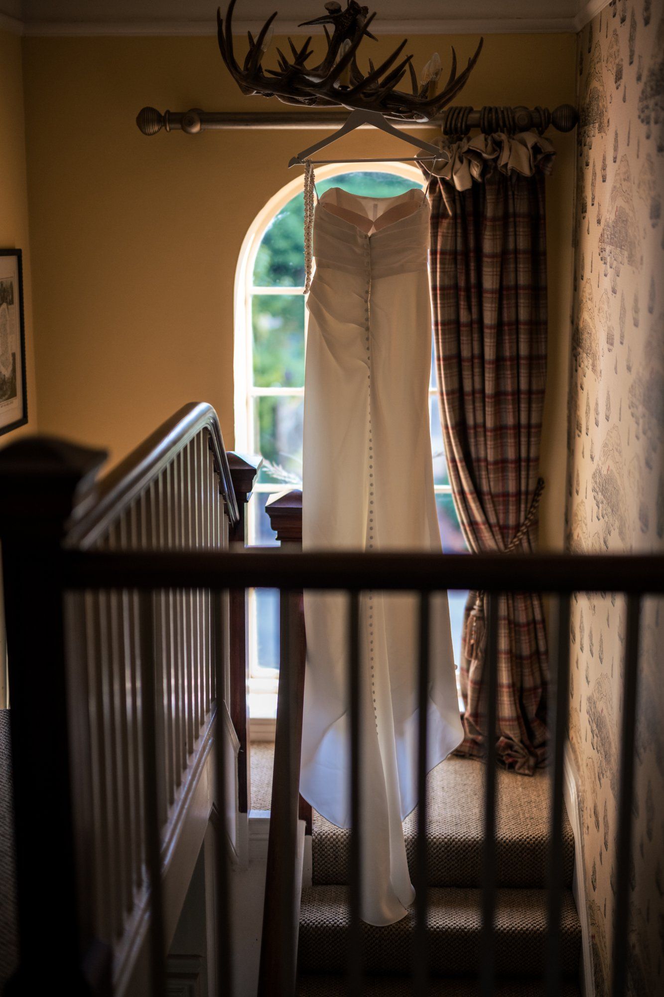 White wedding dress hanging in arched window on staircase landing with antler chandelier and plaid curtains.