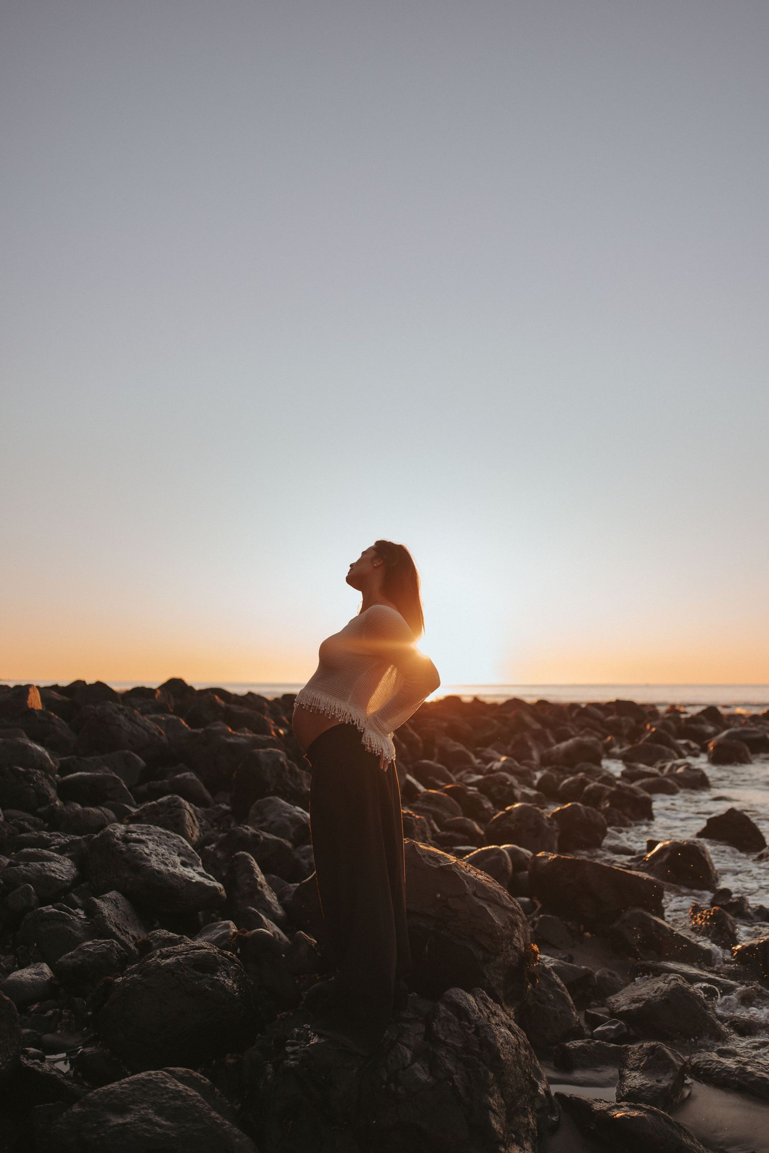 Woman standing on rocky shore at golden sunset, silhouetted against warm orange sky near ocean.