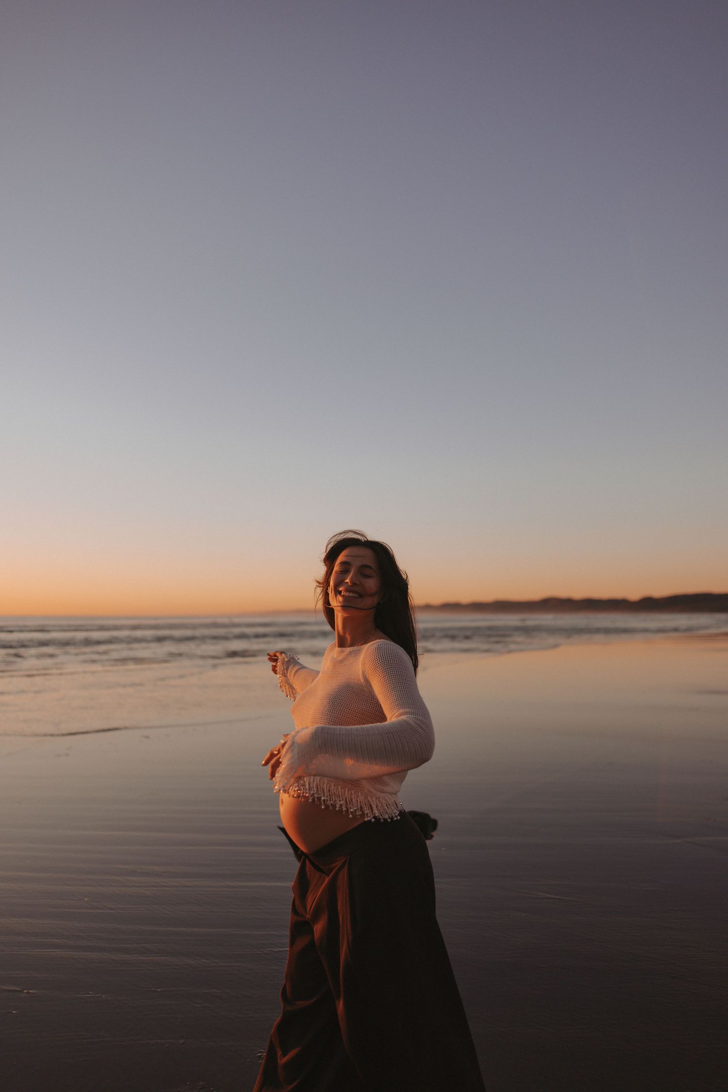 Smiling pregnant woman in white top and dark skirt standing on a beach at sunset with golden sky.
