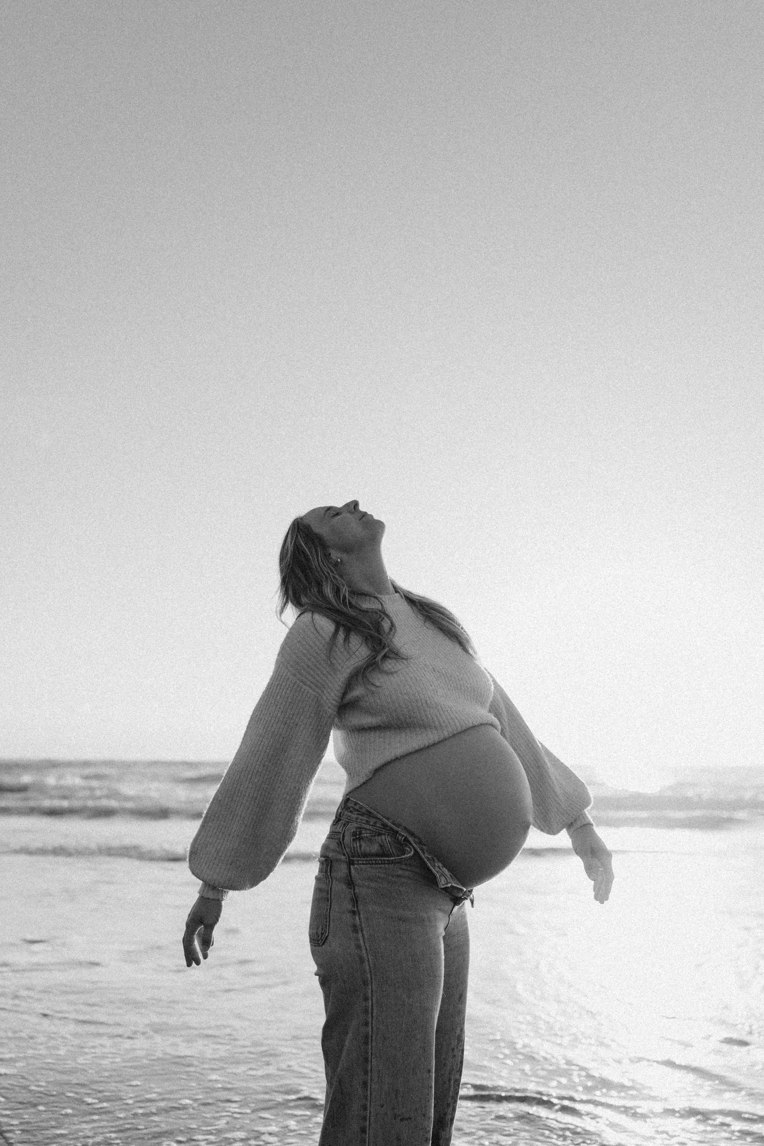Black and white photo of a pregnant woman at the beach in Waikato NZ, head tilted back, arms open wide, wearing a sweater and jeans.