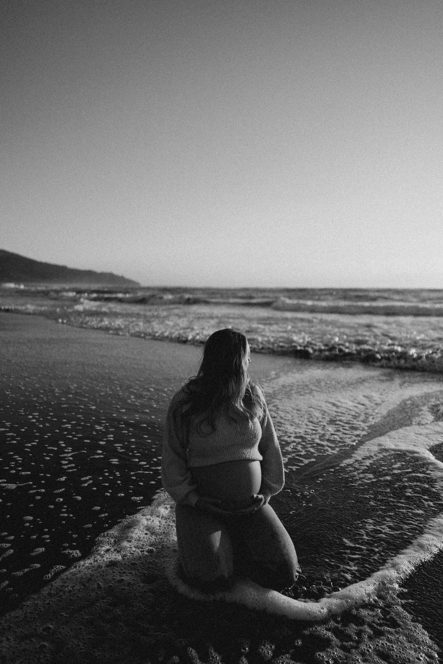Black and white photo of a pregnant woman kneeling on a beach, cradling her belly as ocean waves wash around her.