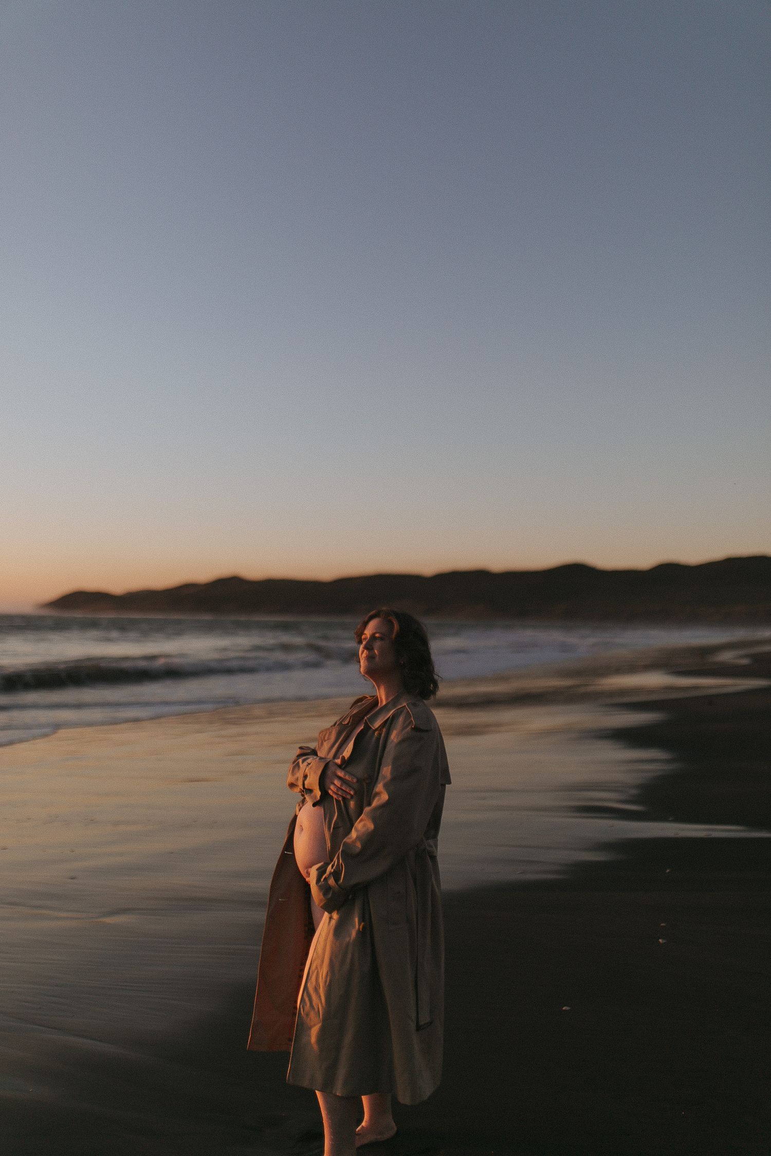 Pregnant woman in trench coat standing barefoot on a beach at sunset with ocean waves and hills in background.