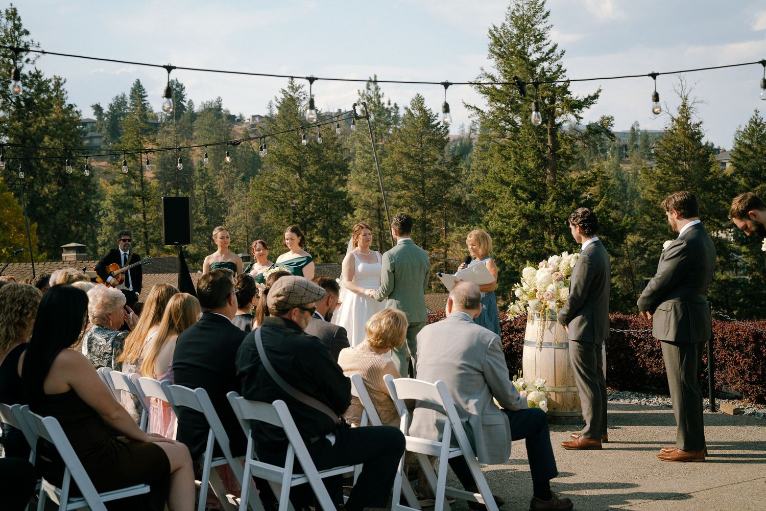 Outdoor wedding ceremony on a rooftop patio with white chairs, floral arrangements, and pine trees in the background.
