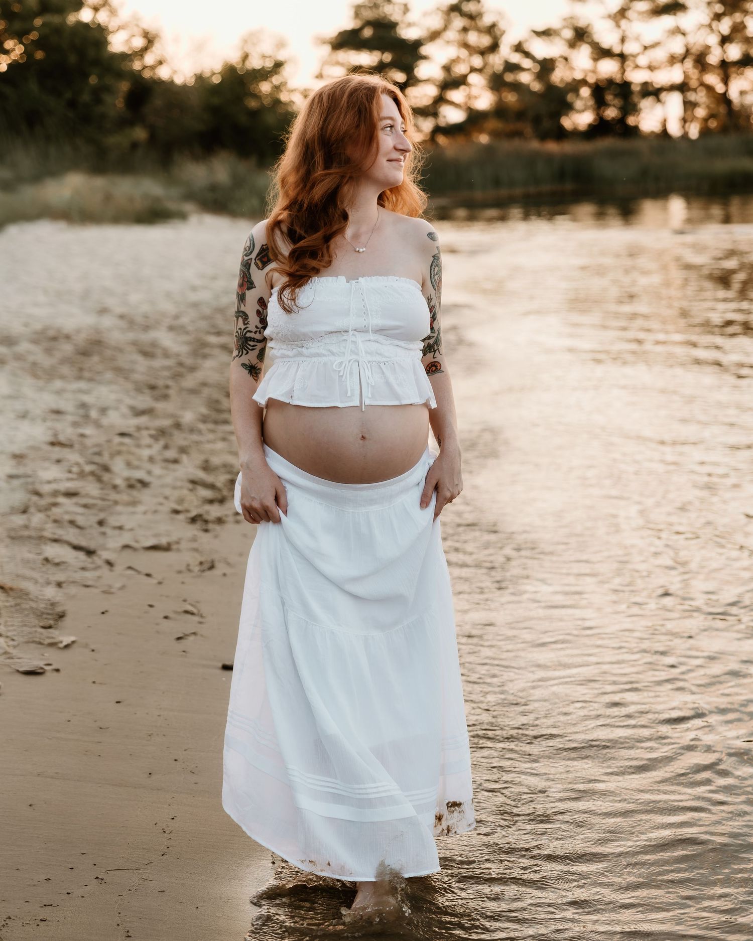 Pregnant woman with red hair in white crop top and maxi skirt standing in shallow water at golden hour.