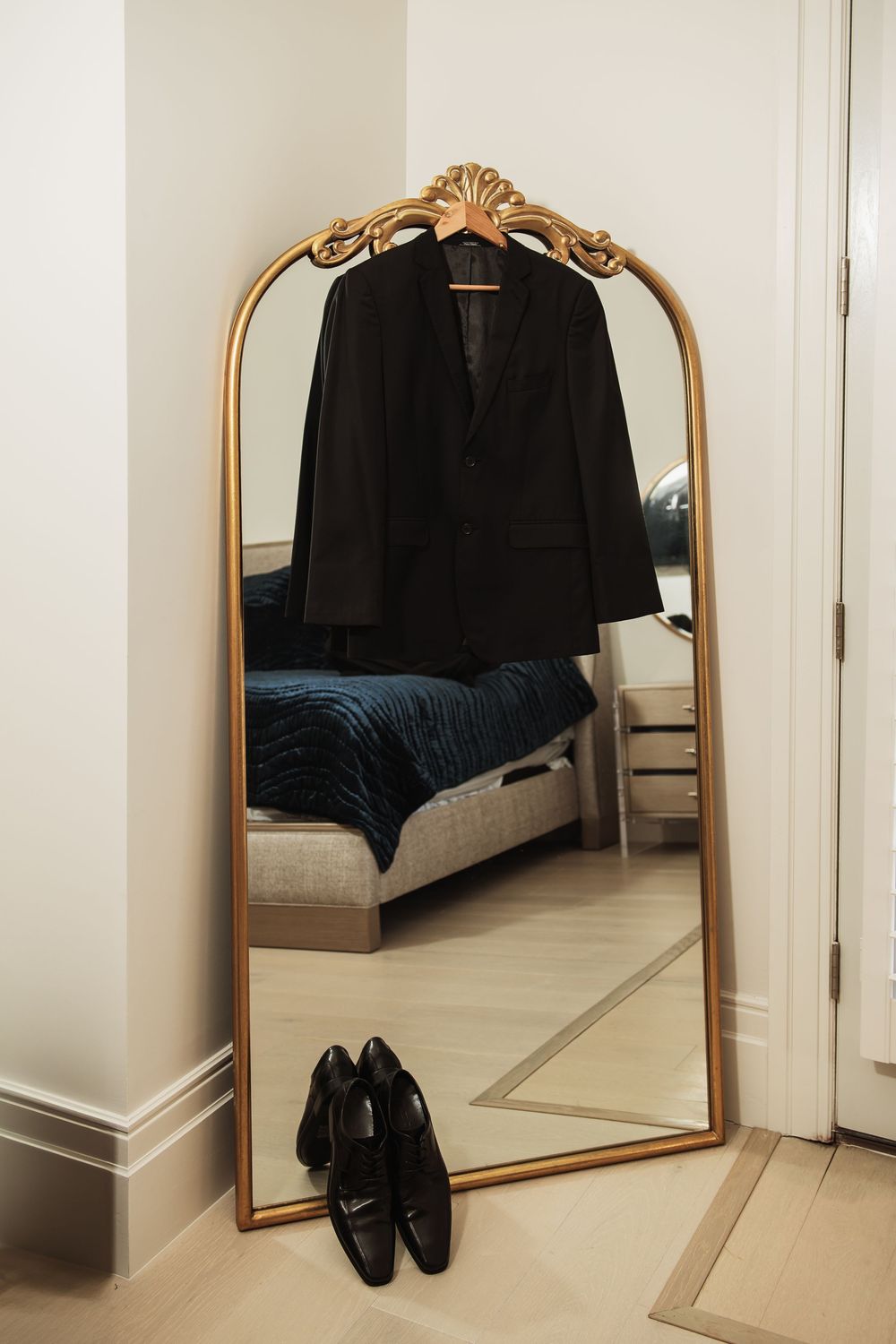 Black suit jacket and dress shoes reflected in ornate gold-framed mirror in elegant bedroom during wedding preparations.