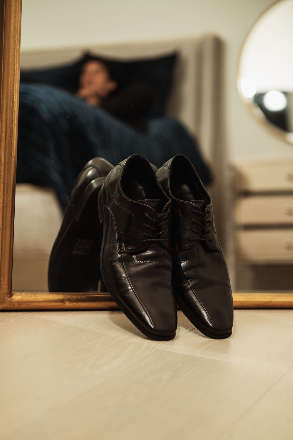 Black leather dress shoes leaning against a mirror reflecting a man resting on a bed in a warmly lit room.