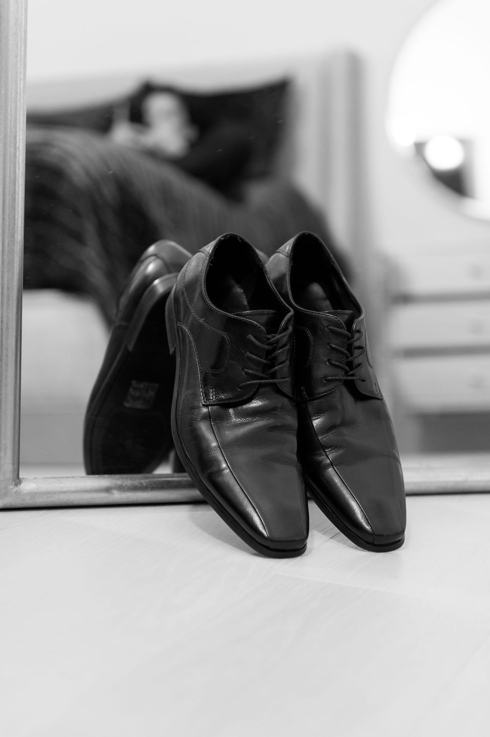 Classic black leather lace-up dress shoes positioned against a mirror in a black and white wedding preparation photo.