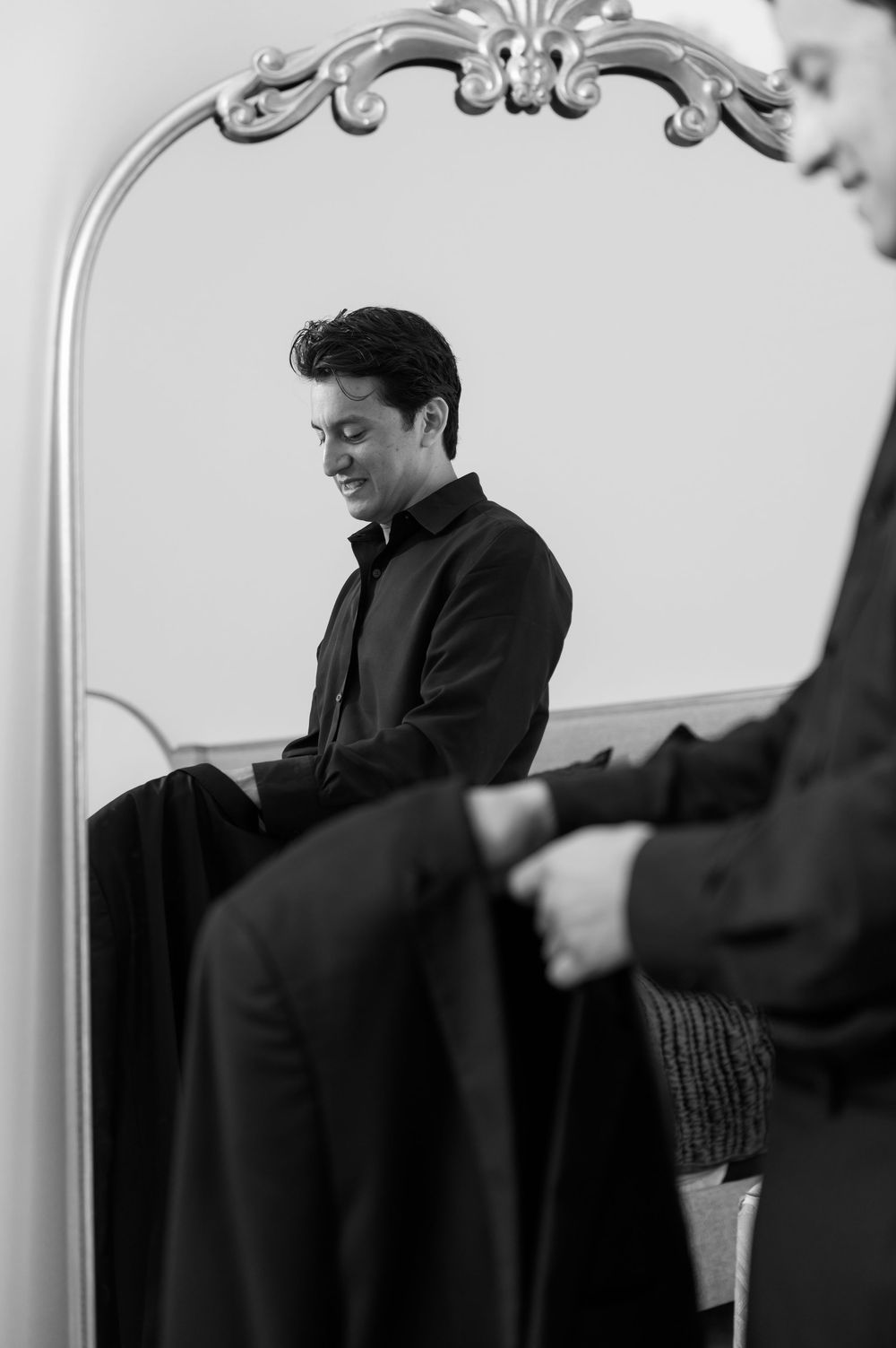 Groom smiling while putting on his black suit jacket reflected in an ornate silver-framed mirror.