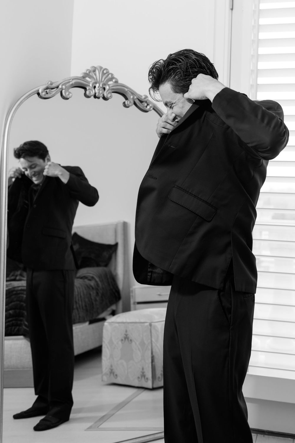 Groom in black suit adjusts collar while viewing reflection in ornate mirror during wedding preparations.