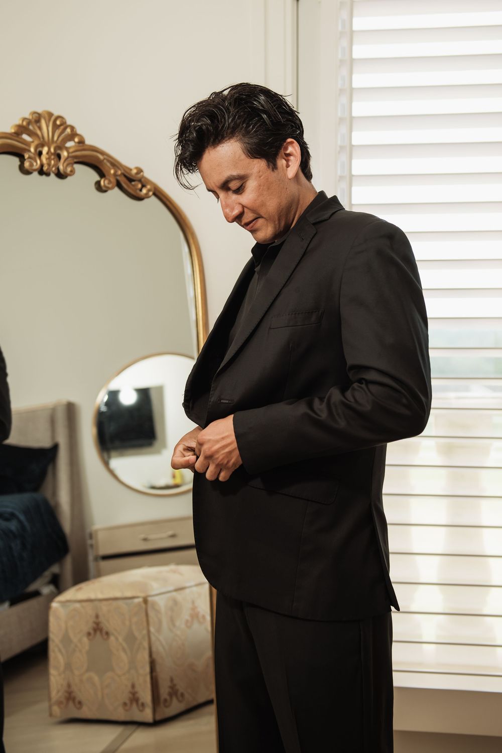 Groom in black suit buttoning jacket before wedding ceremony in elegant room with ornate mirror.