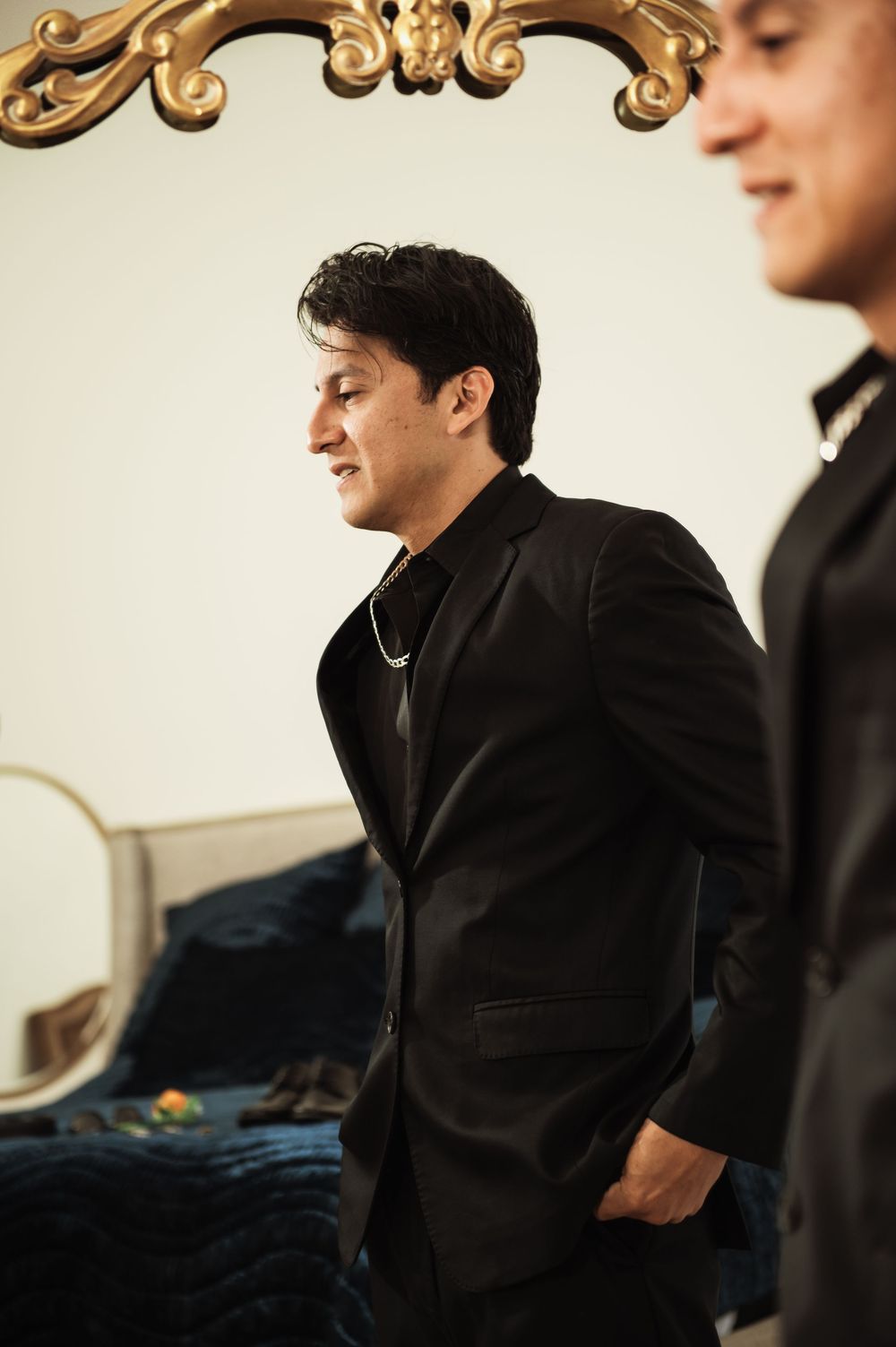 Groom in black suit stands confidently in ornate room with gold-framed mirror during wedding preparations.