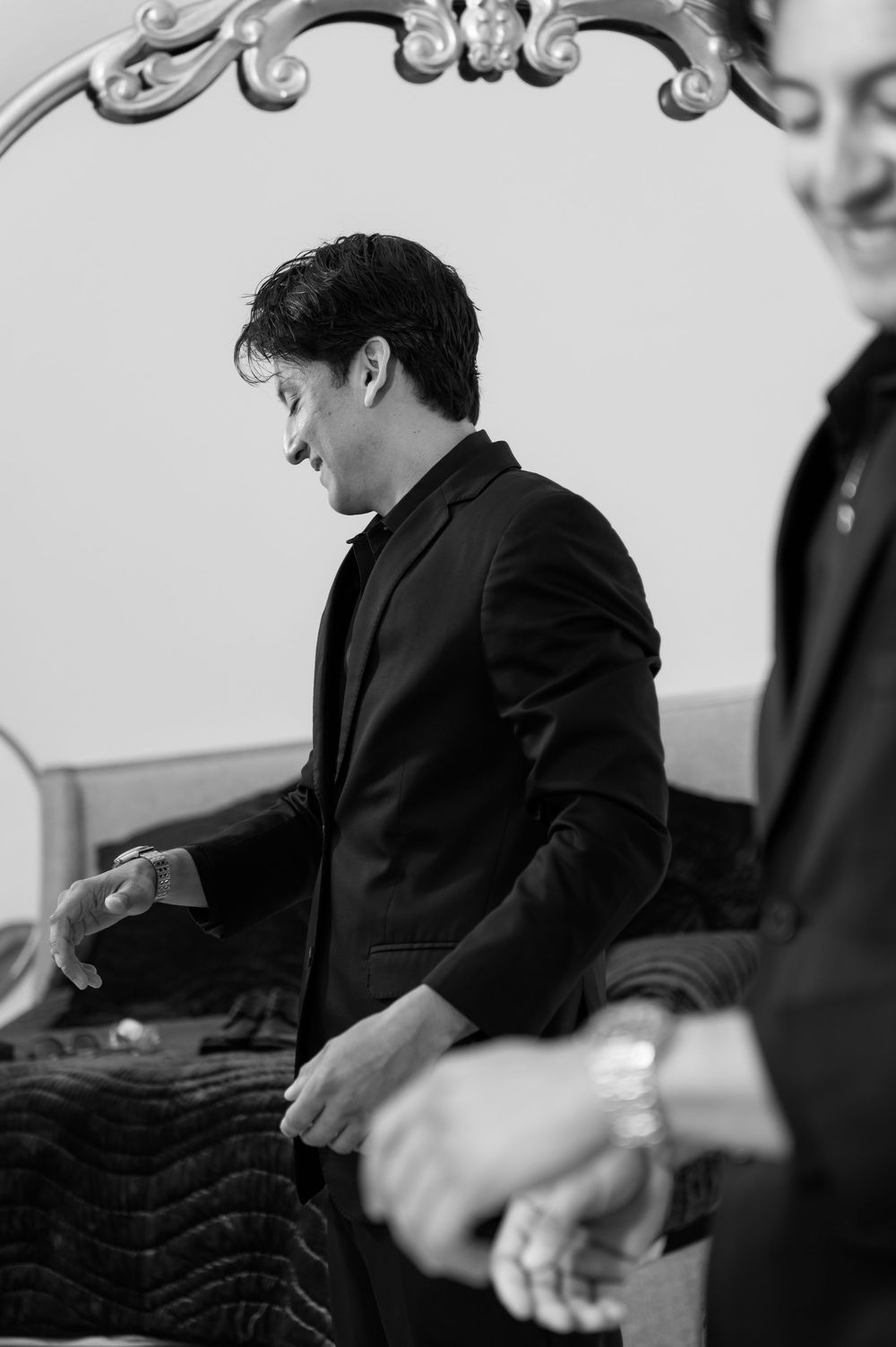 Groom in black suit adjusts jacket in black and white photo during wedding day preparations near ornate mirror.