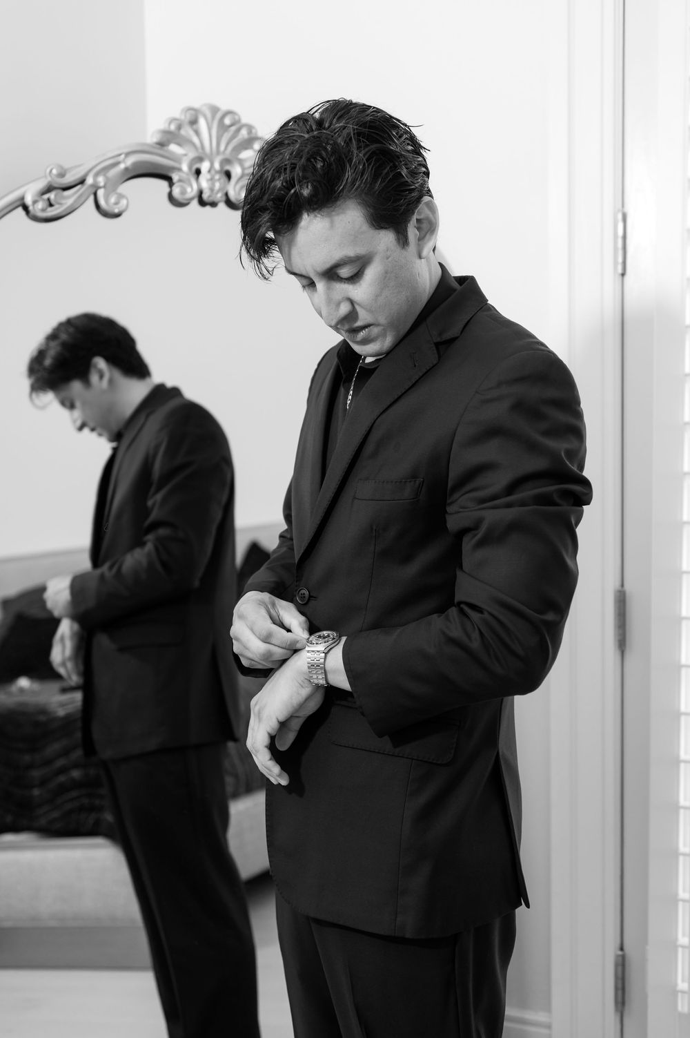 Groom in black suit adjusting watch before wedding ceremony, reflected in ornate mirror.