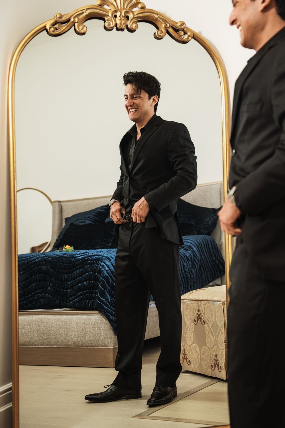 Smiling groom in black suit adjusts jacket while standing before ornate gold mirror in elegant bedroom.