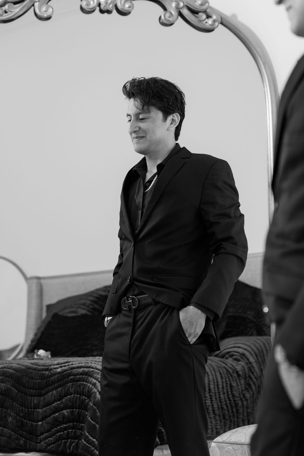 Groom in black suit smiling beside ornate mirror in elegant black and white portrait.
