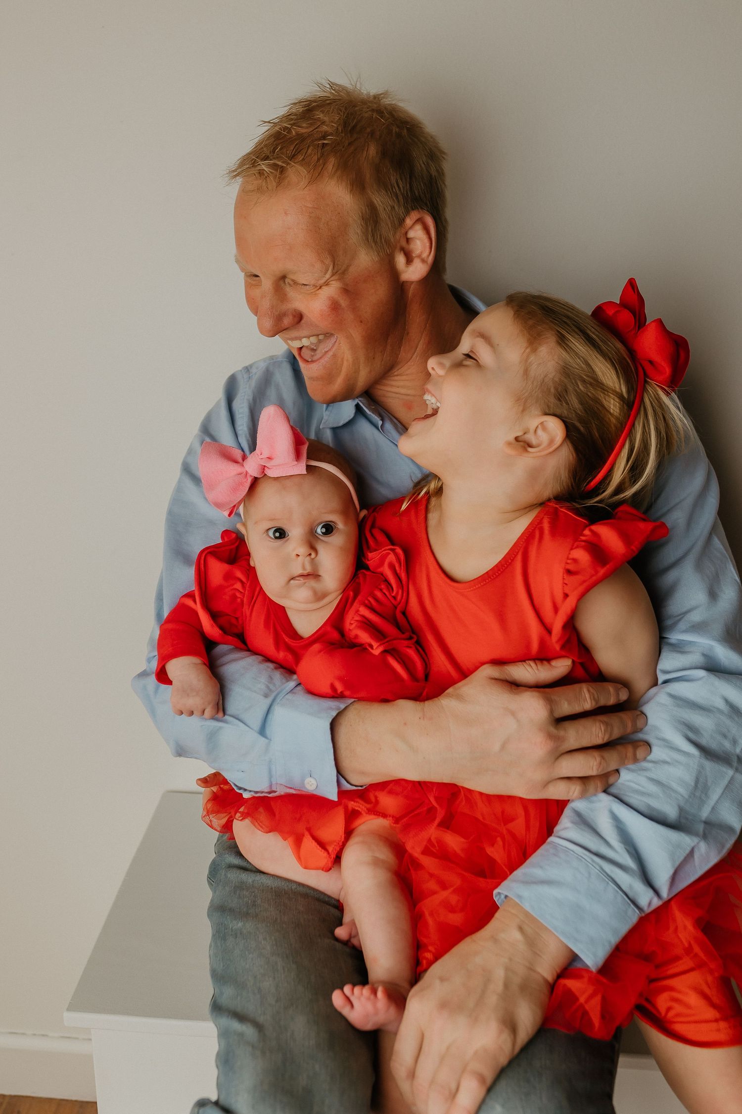 A smiling man holds a baby girl and young girl, both in red dresses, in a warm family portrait.