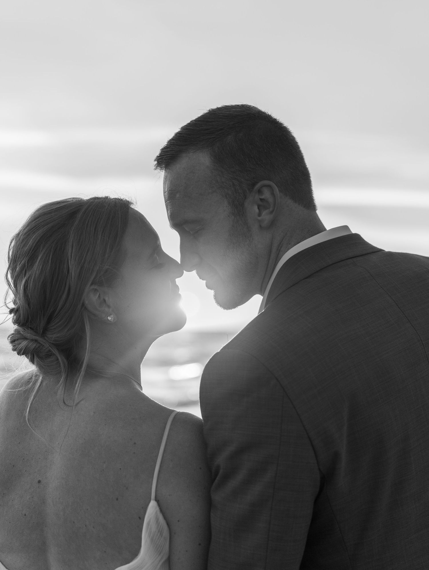 Black and white photo of a couple sharing a romantic moment at sunset on a beach, noses nearly touching.