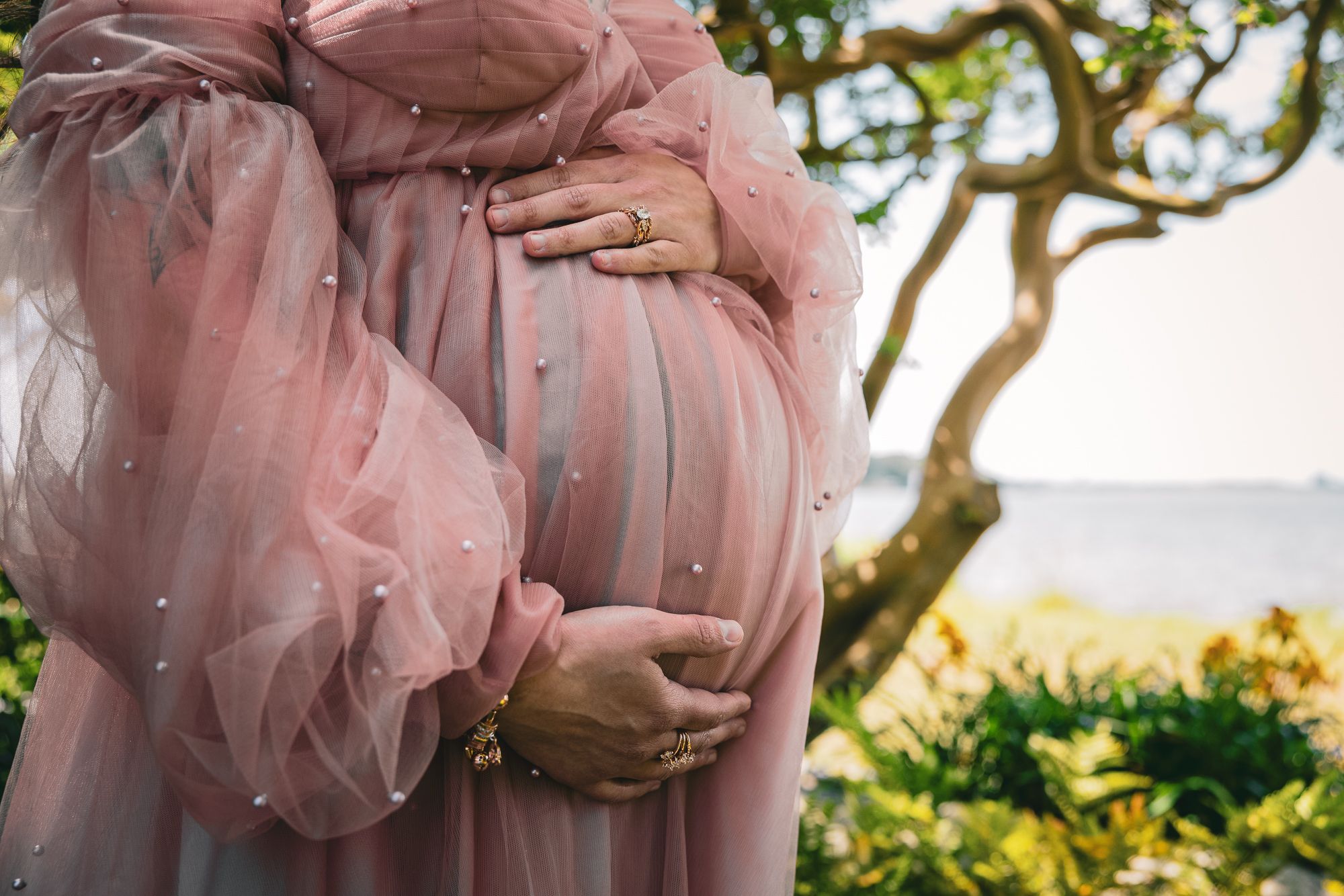 Pregnant woman in a pink tulle maternity gown cradling her baby bump outdoors with greenery in the background.