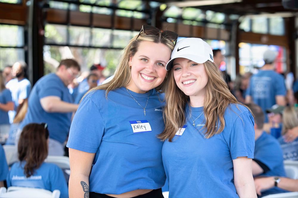 Two smiling women in blue volunteer t-shirts and name tags pose together at an outdoor event.