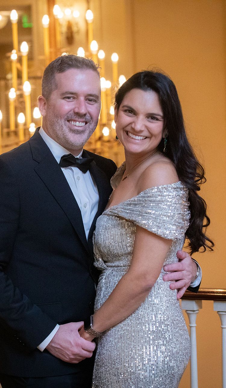 Elegant couple smiling at formal event, man in tuxedo and woman in silver sequin off-shoulder gown, candlelit background.