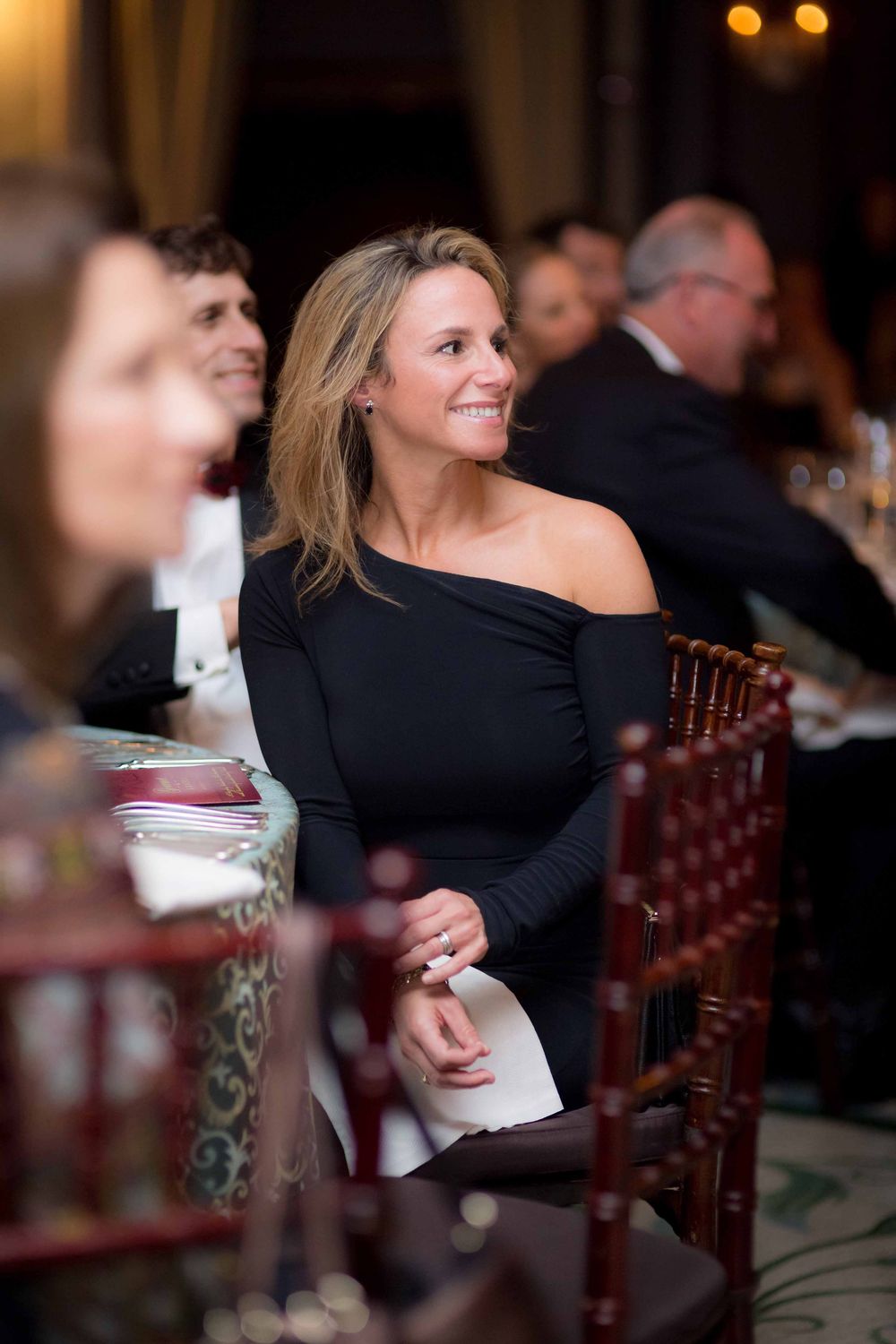 Elegant woman in black off-shoulder dress seated at formal event, smiling gracefully at a gala dinner.