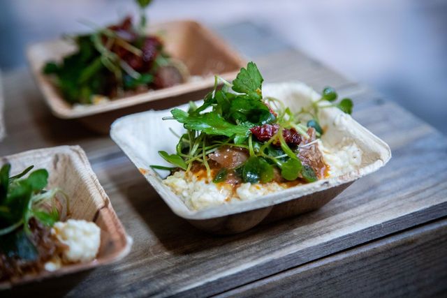 Fresh grain bowl topped with greens, herbs, and savory toppings served in eco-friendly takeout containers on wood.