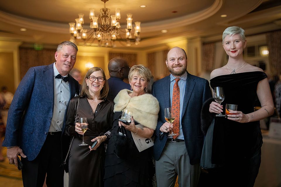 Five elegantly dressed guests smile and hold drinks at a formal gala event in a grand ballroom.