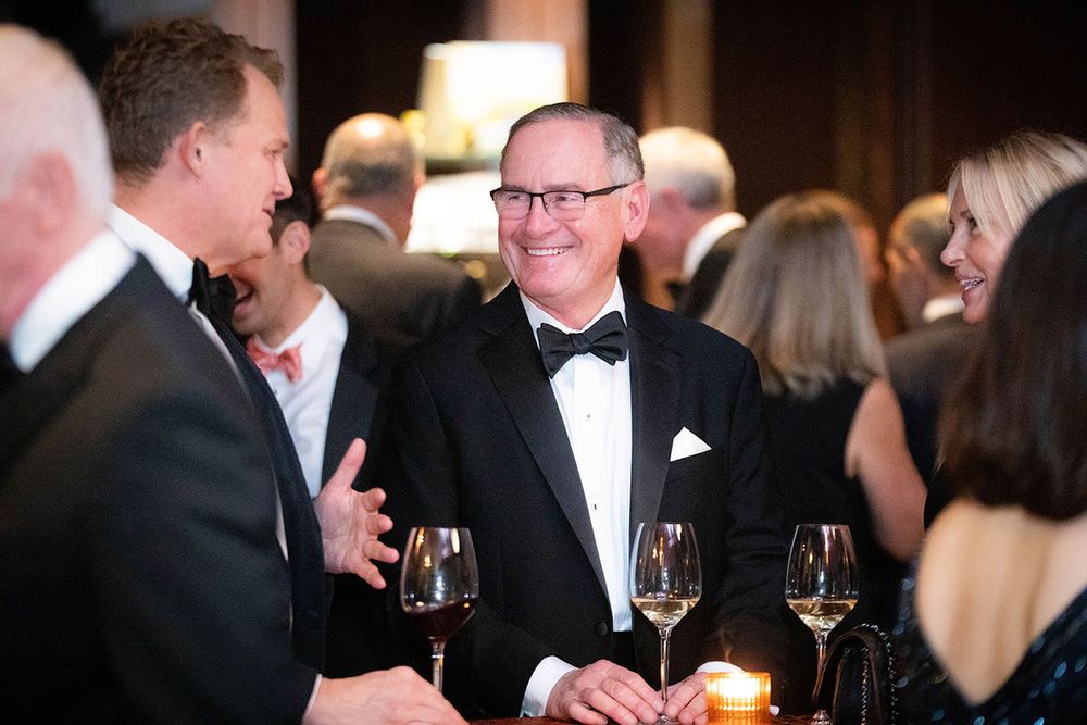 Men in tuxedos socializing at a formal gala event, holding red wine glasses in an elegant venue.