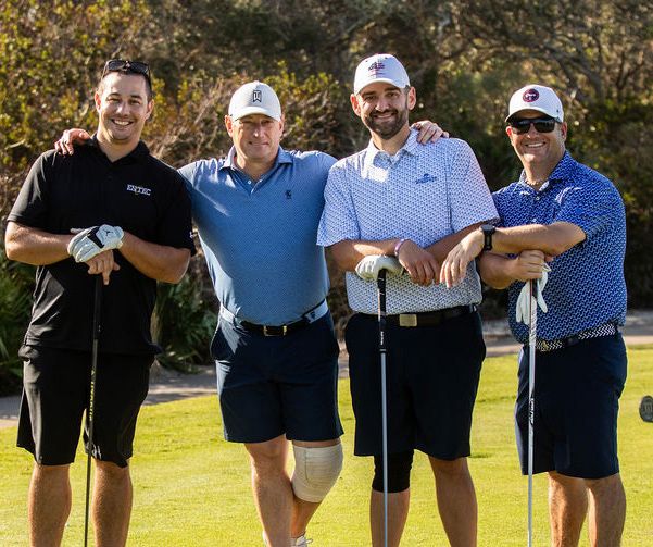 Four men smiling and posing together on a golf course, holding clubs, wearing casual golf attire.