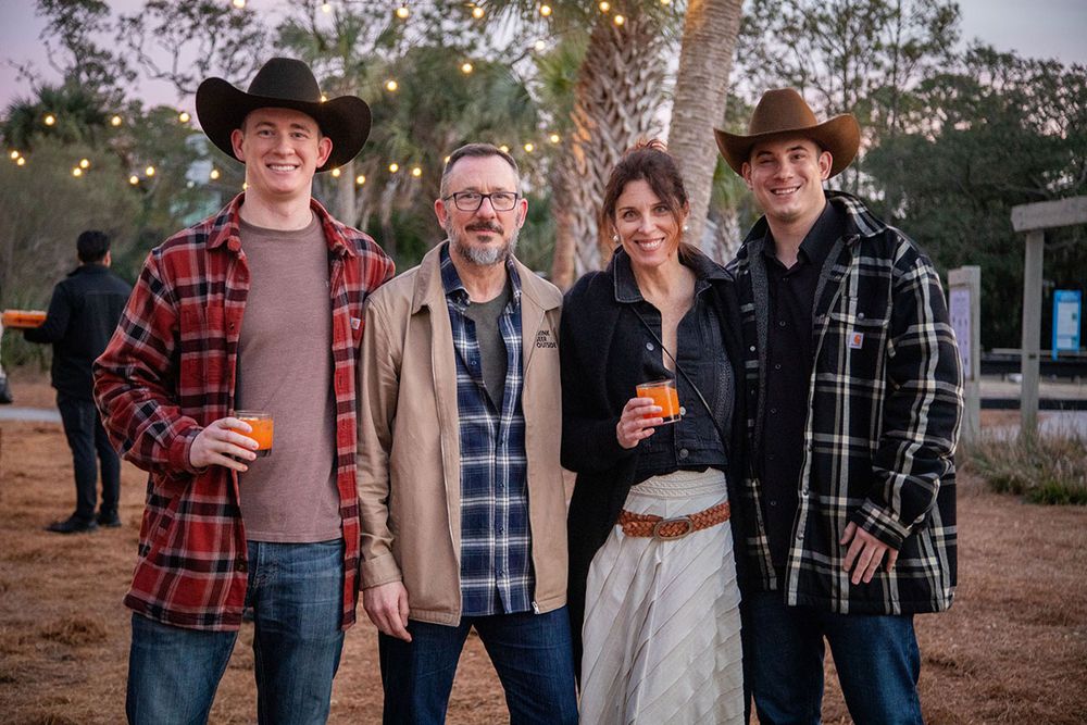 Four friends pose outdoors at a evening event, two wearing cowboy hats, string lights visible in background.