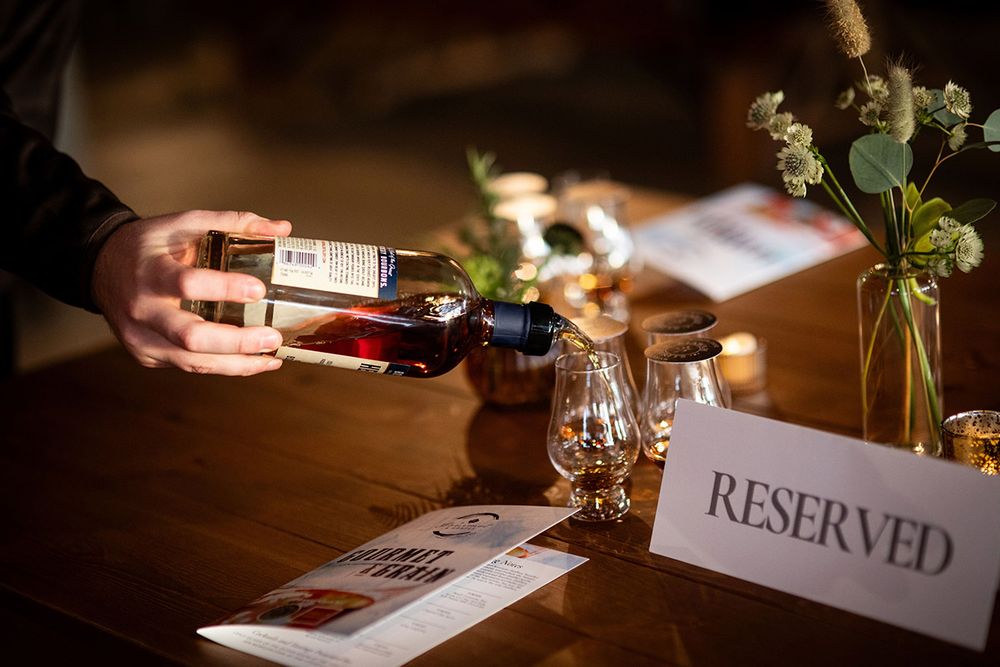 Hand pouring whiskey bottle at reserved table setting with glassware and floral decor in dimly lit venue.