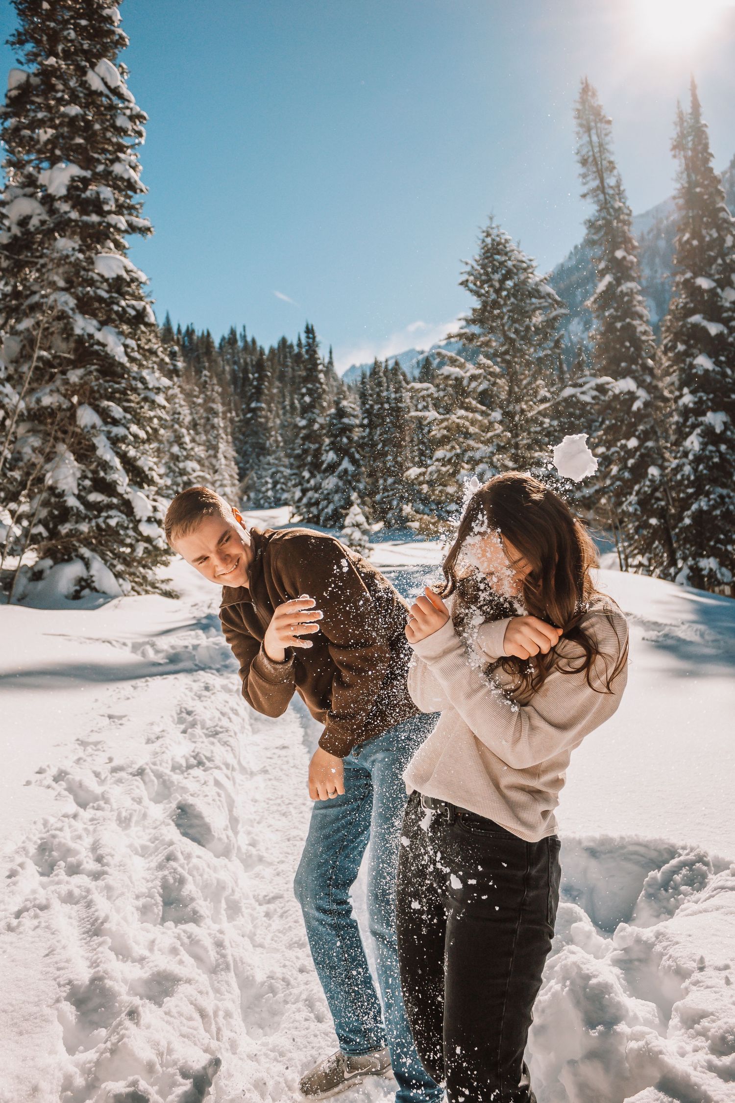 Couple having a snowball fight in a snowy winter forest with tall pine trees and bright blue sky.