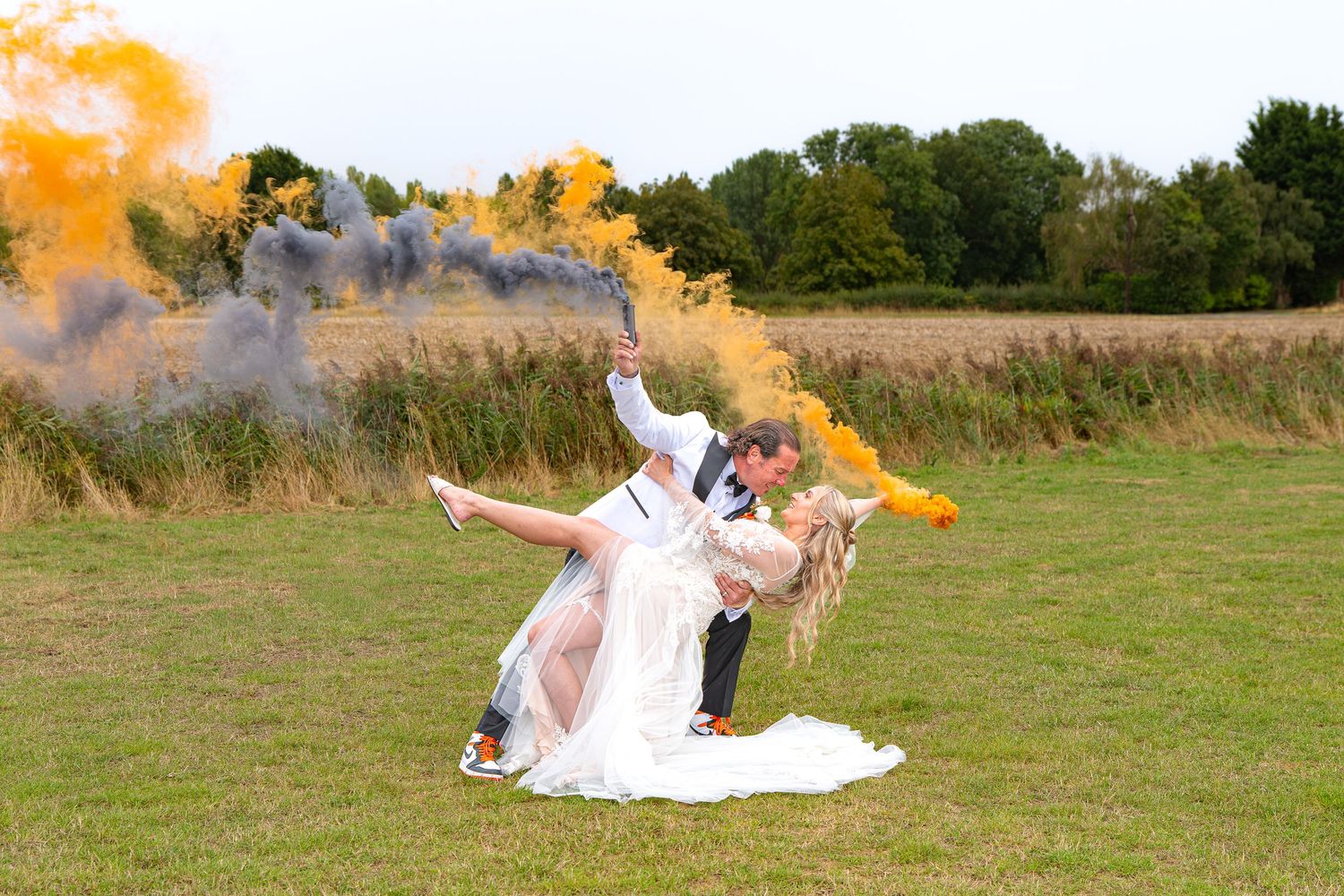 Bride and groom share a romantic dip kiss while holding colorful orange smoke bombs during outdoor wedding photo shoot.