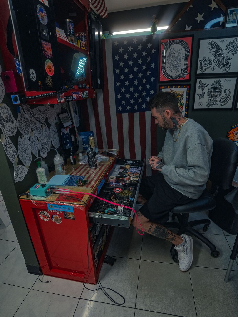 Tattoo artist Karl Cooper preps a machine at his station at Sacred Art Tattoo in Honolulu, Hawaii, flags, flash art & ink tools around him.