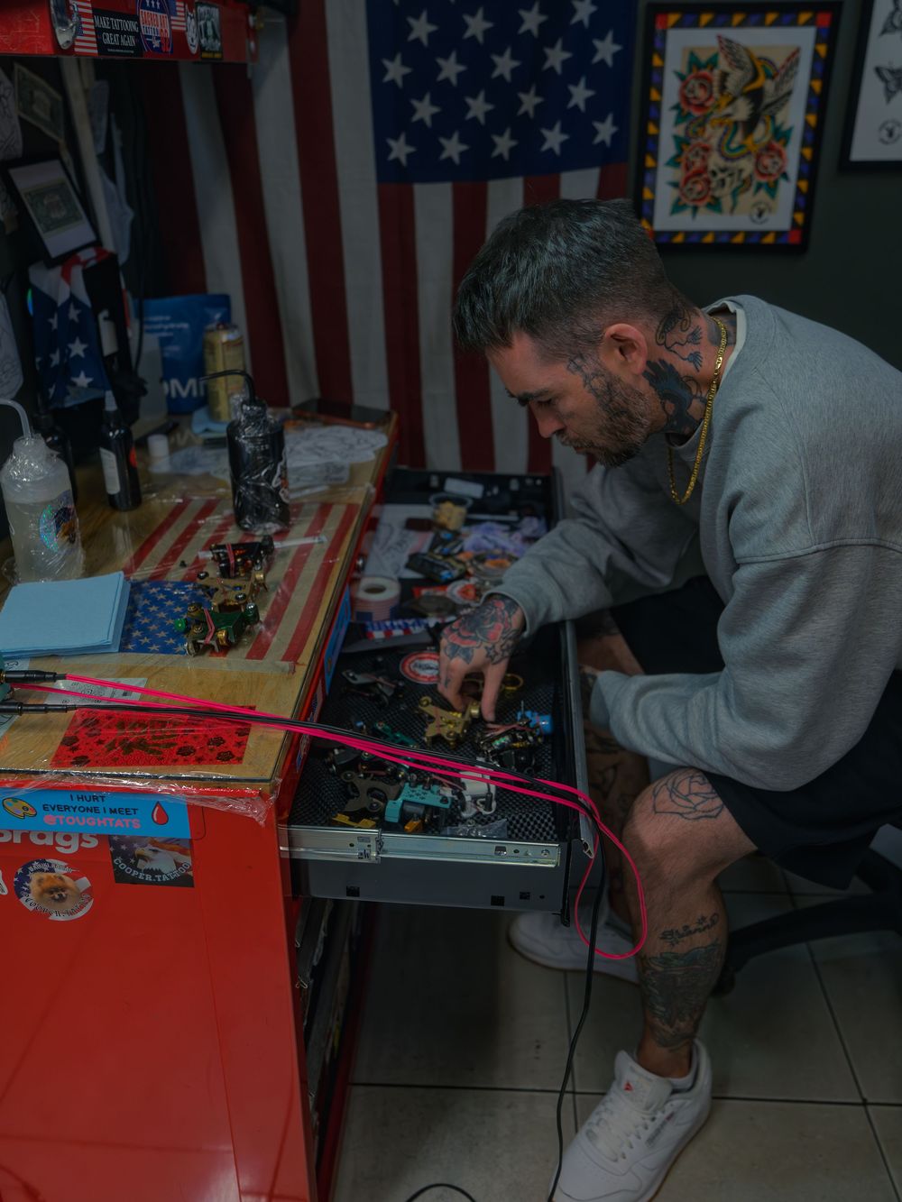 Tattoo artist Karl Cooper picks a coil machine from a drawer full of them at Sacred Art Tattoo in Honolulu, Hawaii, flags and flash behind.