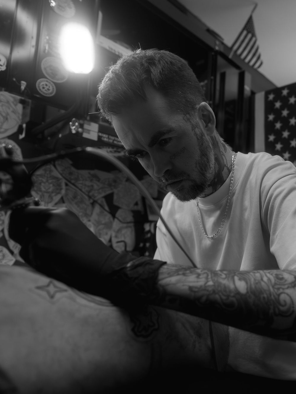 Black-and-white portrait of tattoo artist Karl Cooper working intently on a leg tattoo at Sacred Art Tattoo in Honolulu, Hawaii, lamp lit.