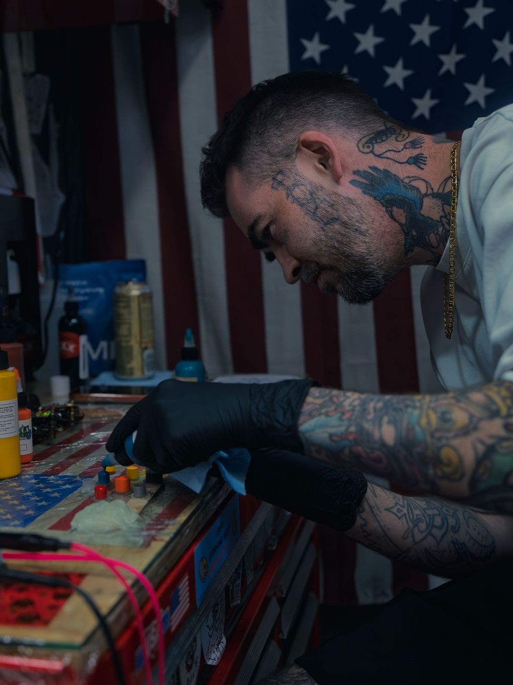 Tattoo artist Karl Cooper leans over his flag-draped station mixing ink caps at Sacred Art Tattoo in Honolulu, Hawaii, tattooed arms sleeved