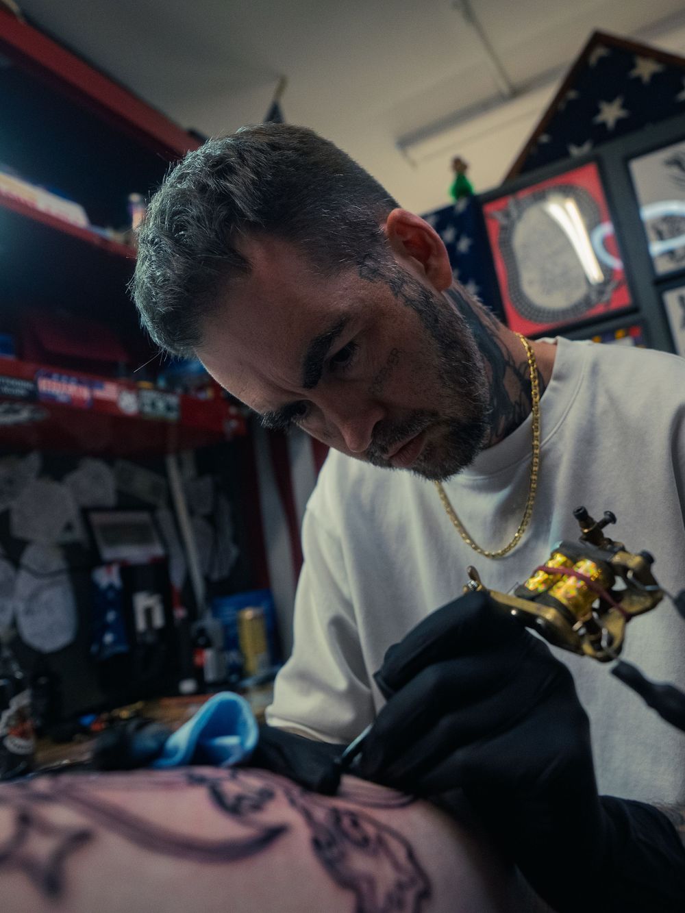 Tattoo artist Karl Cooper focuses on a leg tattoo, brass coil machine in hand, at Sacred Art Tattoo in Honolulu, Hawaii, flag behind him.