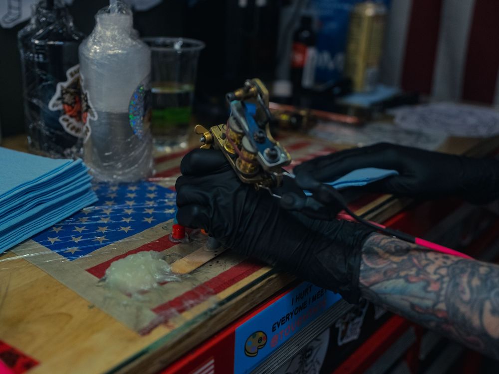 Karl Cooper's gloved, tattooed hand loads a brass tattoo machine at his flag-draped station at Sacred Art Tattoo in Honolulu, Hawaii.