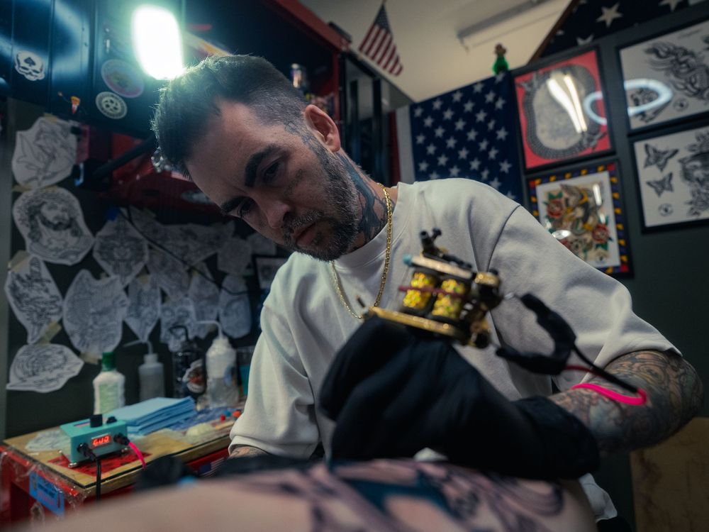 Tattoo artist Karl Cooper leans in with his machine as he works on a client's leg at Sacred Art Tattoo in Honolulu, Hawaii, flash behind.
