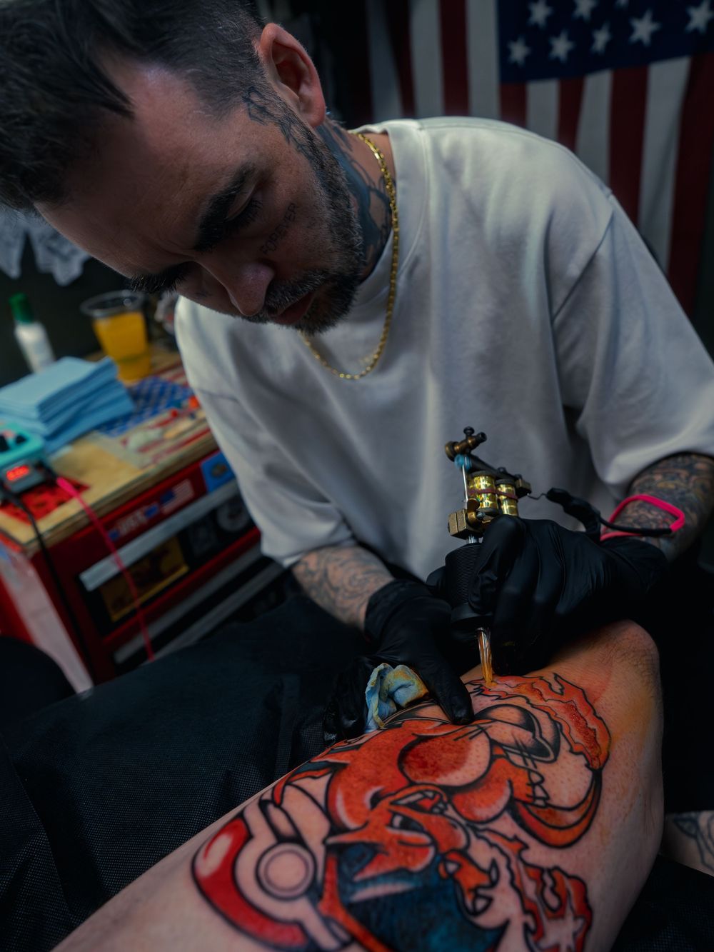 Tattoo artist Karl Cooper leans in with his machine, coloring a bold orange leg tattoo at Sacred Art Tattoo in Honolulu, Hawaii, flag behind