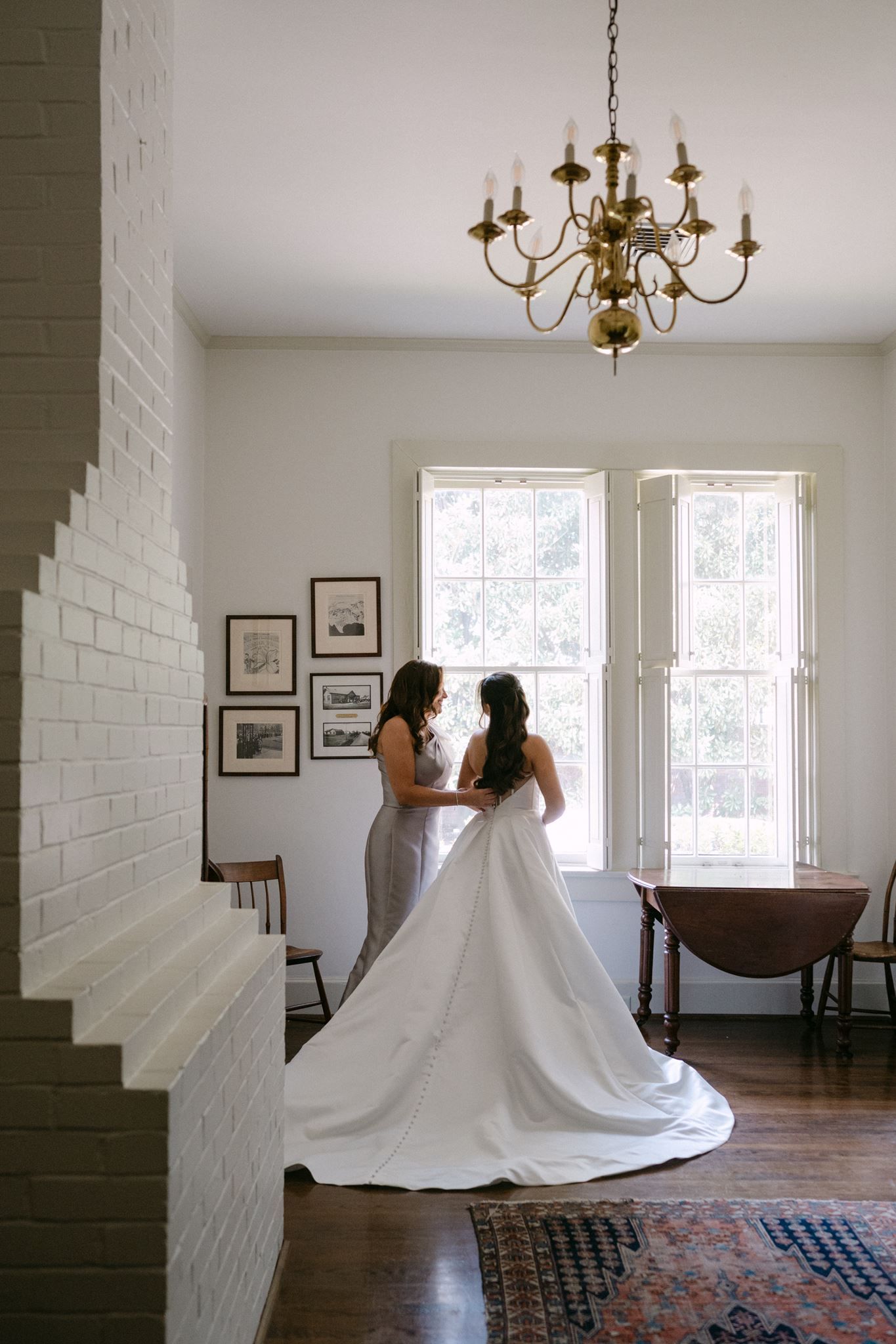 Mother of the bride buttoning her daughter's dress in front of a large window.
