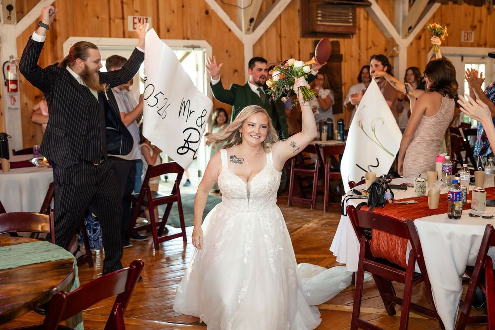 Bride in white gown celebrates grand entrance at rustic barn wedding reception with cheering guests.